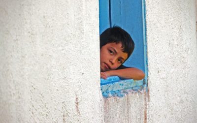 A young boy rest his head and arm on a window sill and peeks out of a house.