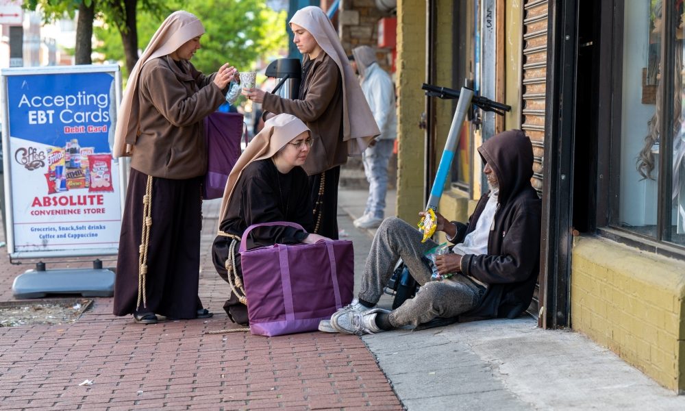 Sister Giovana of the Most Holy Name of Jesus (squatting) joins Sister Samaritan of Scourged Love (left) and Sister Maria Clara of the Crucified in giving food to a man near Lexington Market in Baltimore May 3, 2023. (OSV News/Kevin J. Parks, Catholic Review)