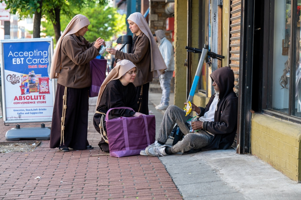 Sister Giovana of the Most Holy Name of Jesus (squatting) joins Sister Samaritan of Scourged Love (left) and Sister Maria Clara of the Crucified in giving food to a man near Lexington Market in Baltimore May 3, 2023. (OSV News/Kevin J. Parks, Catholic Review)