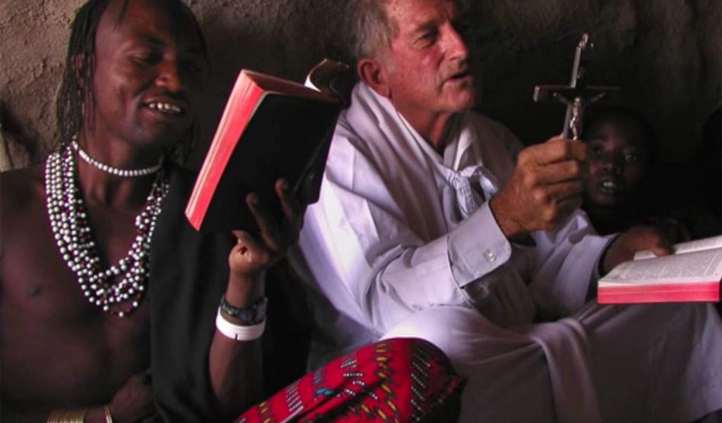 A Maryknoll priest sits on the floor holding a crucifix and a Bible, praying with a small group of people.
