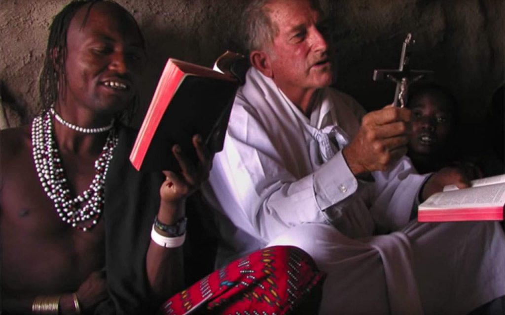 A Maryknoll priest sits on the floor holding a crucifix and a Bible, praying with a small group of people.