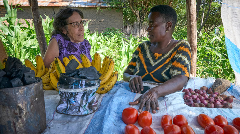 Maryknoll Lay Missioner Joanne Miya (left) talks with Laurencia Makanya, who sells vegetables and charcoal from a small stall in front of her home in a village near Mwanza, Tanzania. Makanya, who is HIV positive, participates in a savings group coordinated by Uzima Centre, where Miya served as director from 2006 until January 2026. (Paul Jeffrey/Tanzania)