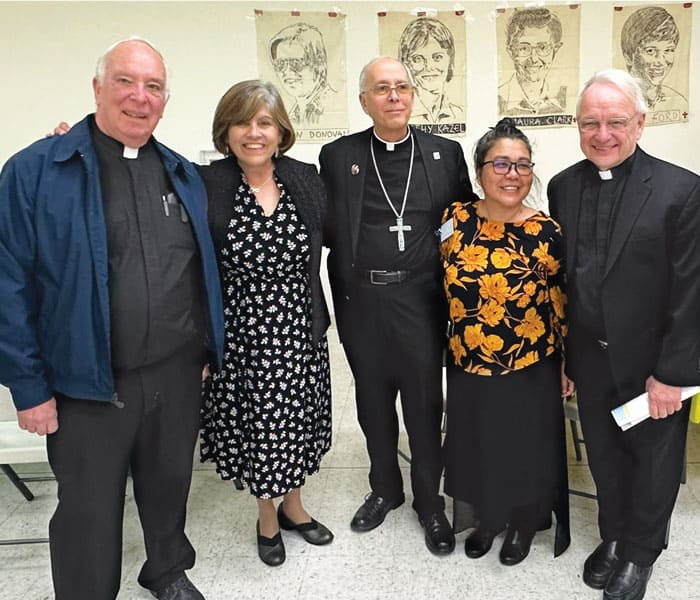 De Izq. a dcha.: El Padre Maryknoll Finch, Ramírez, el Obispo Mark Seitz de El Paso, la Hermana Maryknoll Montiel y el Padre Nadeau posan frente a los retratos de las cuatro mártires de El Salvador. (Cortesía de los Misioneros Laicos Maryknoll)