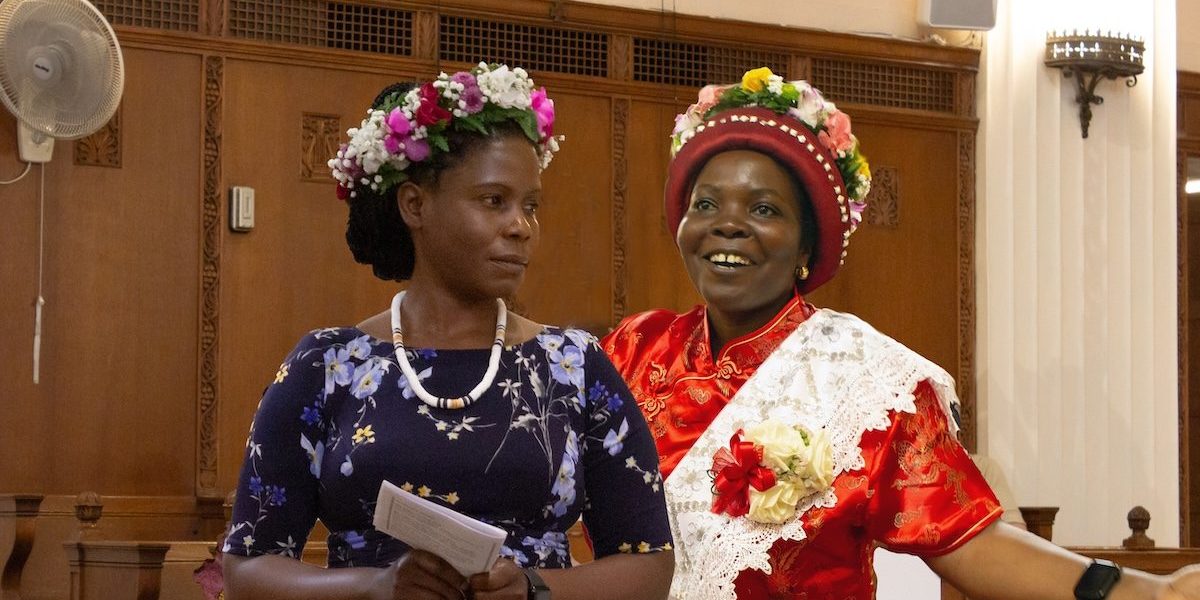 Dos mujeres con conjuntos coloridos y sombreros están de pie en una iglesia.
