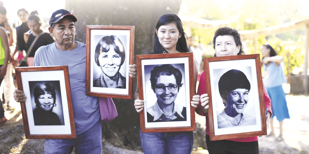 Un hombre y dos mujeres sostienen cuatro grandes fotos enmarcadas en blanco y negro de rostros de personas.