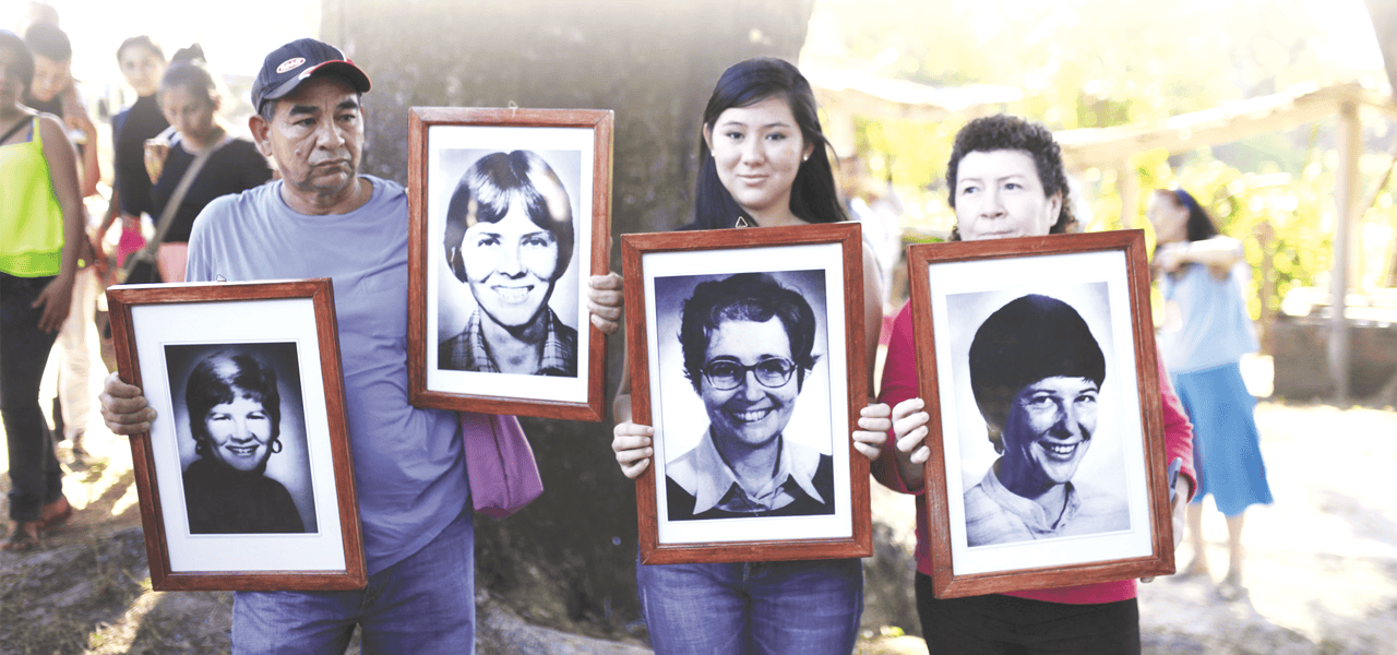 Un hombre y dos mujeres sostienen cuatro grandes fotos enmarcadas en blanco y negro de rostros de personas.