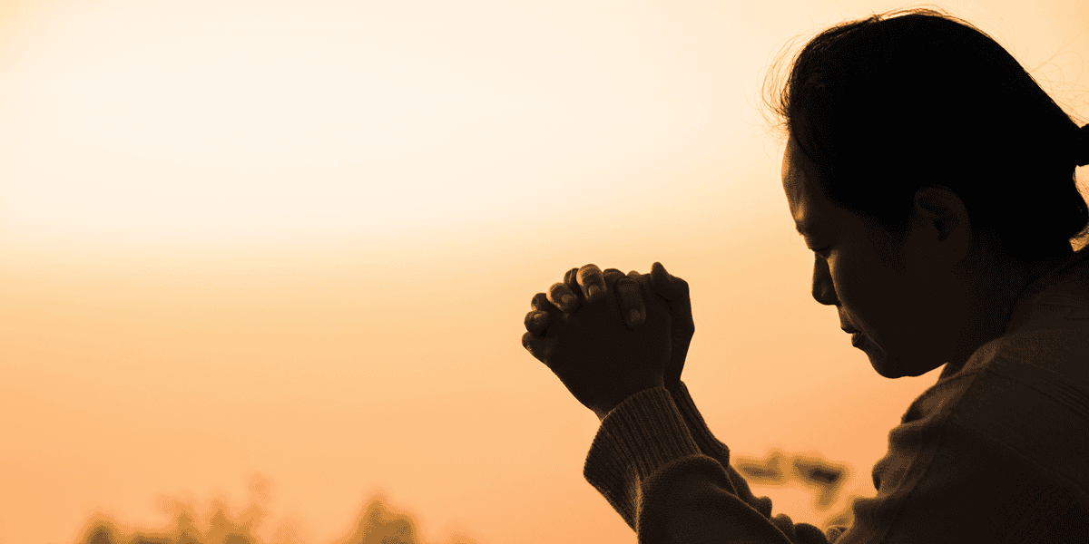 Silueta de una mujer orando a Dios con un atardecer de fondo.
