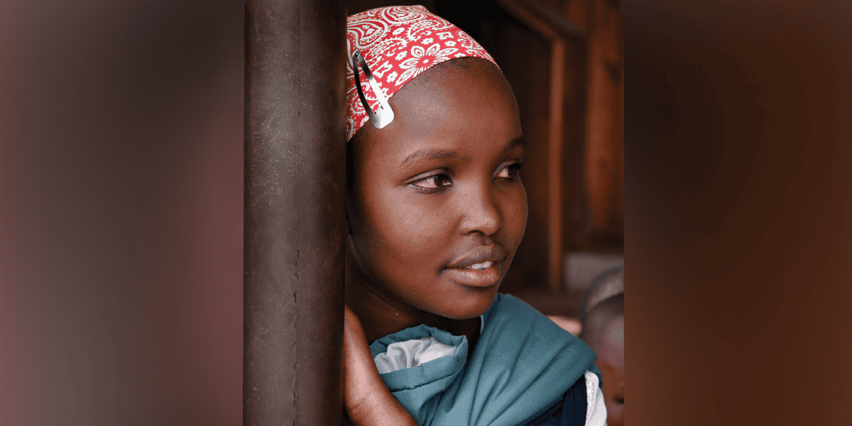 Una niña pequeña que lleva un pañuelo rojo mira hacia un lado mientras se inclina contra un poste.
