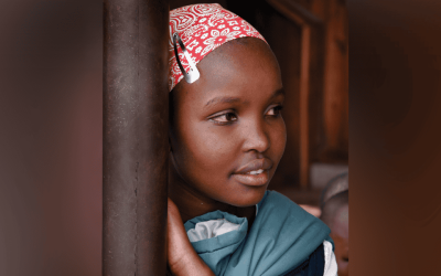 Una niña pequeña que lleva un pañuelo rojo mira hacia un lado mientras se inclina contra un poste.