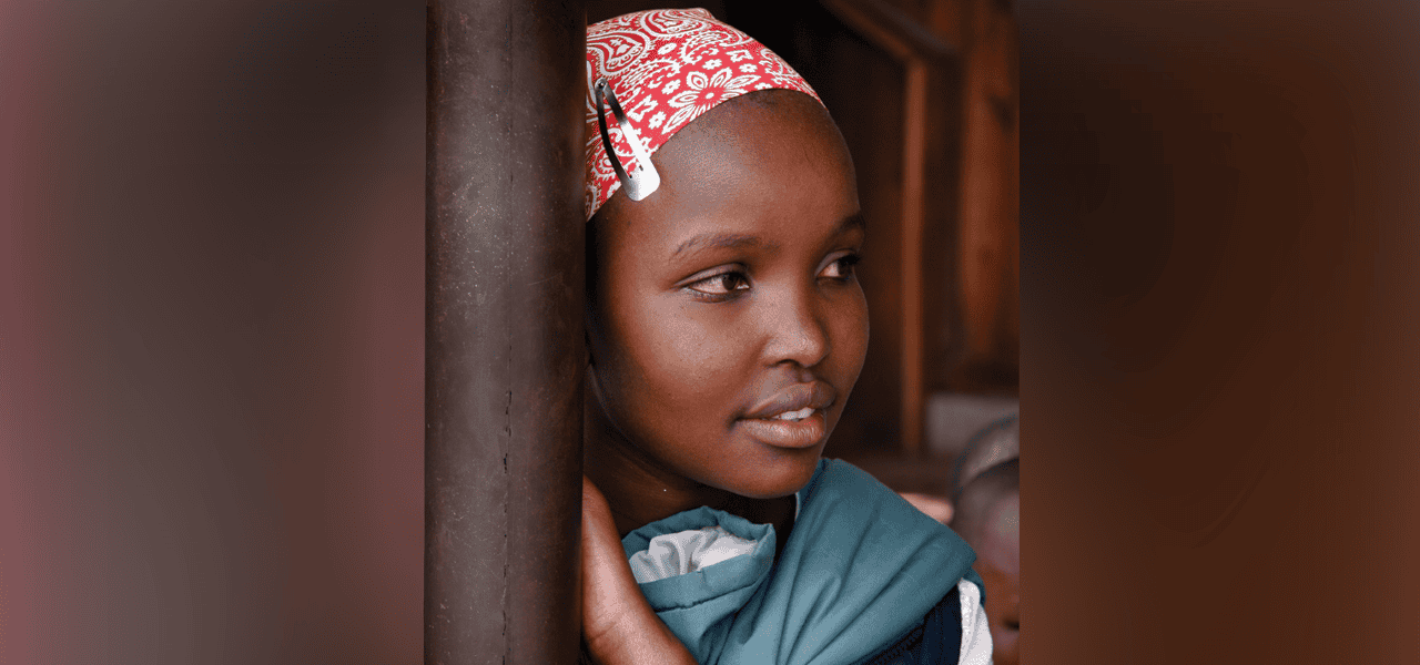 Una niña pequeña que lleva un pañuelo rojo mira hacia un lado mientras se inclina contra un poste.