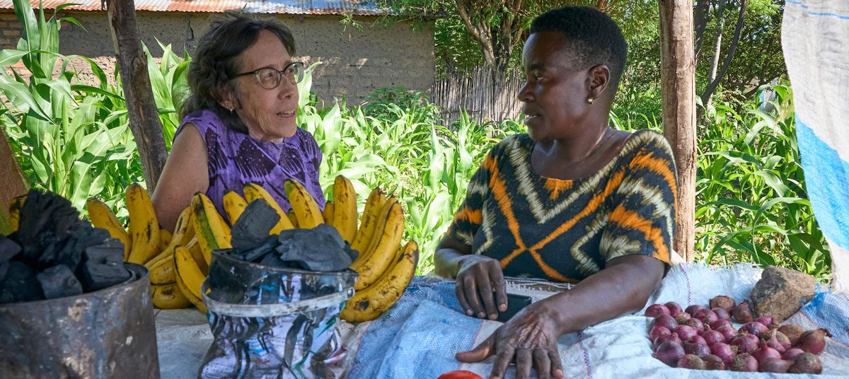 La misionera laica Maryknoll Joanne Miya (izquierda) habla con Laurencia Makanya, quien vende verduras y carbón vegetal en un pequeño puesto frente a su casa, en una aldea cercana a Mwanza, Tanzania. Makanya, que es seropositiva, participa en un grupo de ahorro coordinado por el Centro Uzima, donde Miya fue directora desde 2006 hasta enero de 2026. (Paul Jeffrey/Tanzania)