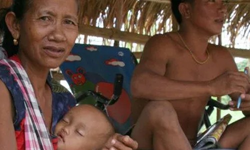 A refugee woman from Burma is sitting in a grass shelter holding her sleeping baby in her arms
