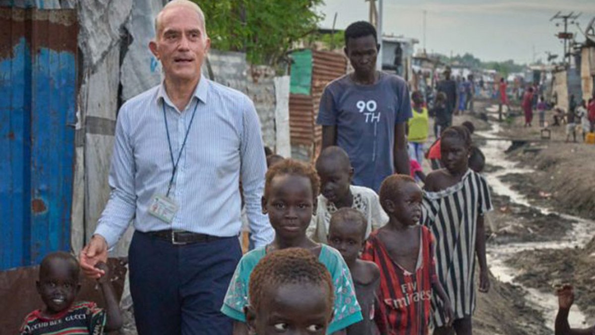 Maryknoll Priest, Fr. Michael Bassano walks with children on a dirt road through makeshift shelters