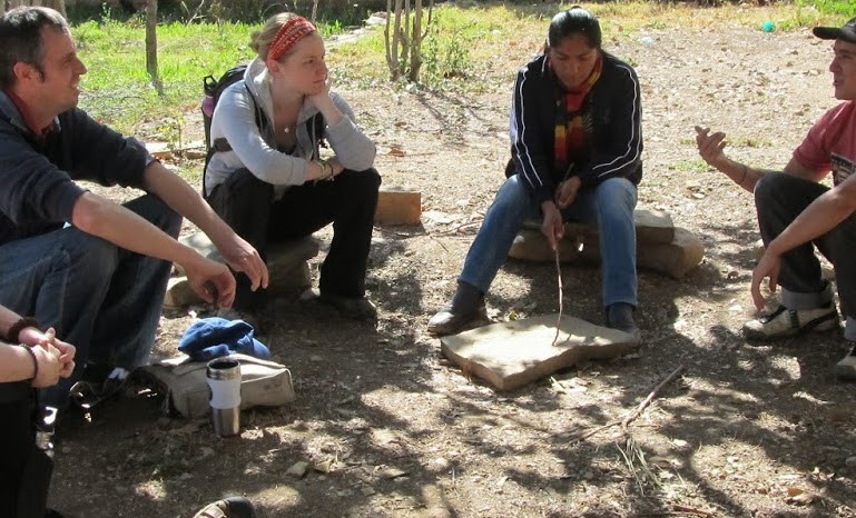 A group of people sitting in a semi-circle under the shade of a tree.