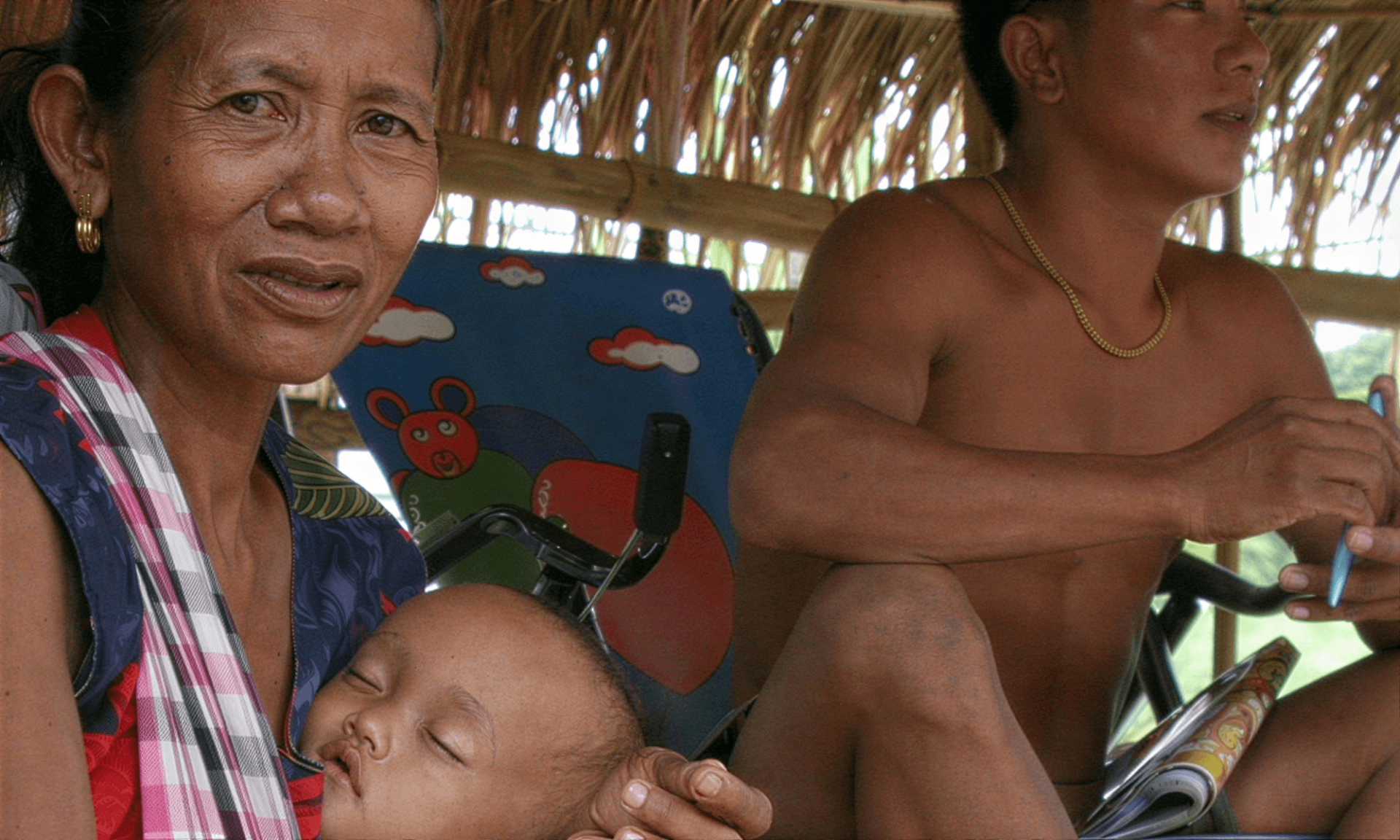 A refugee woman from Burma is sitting in a grass shelter holding her sleeping baby in her arms