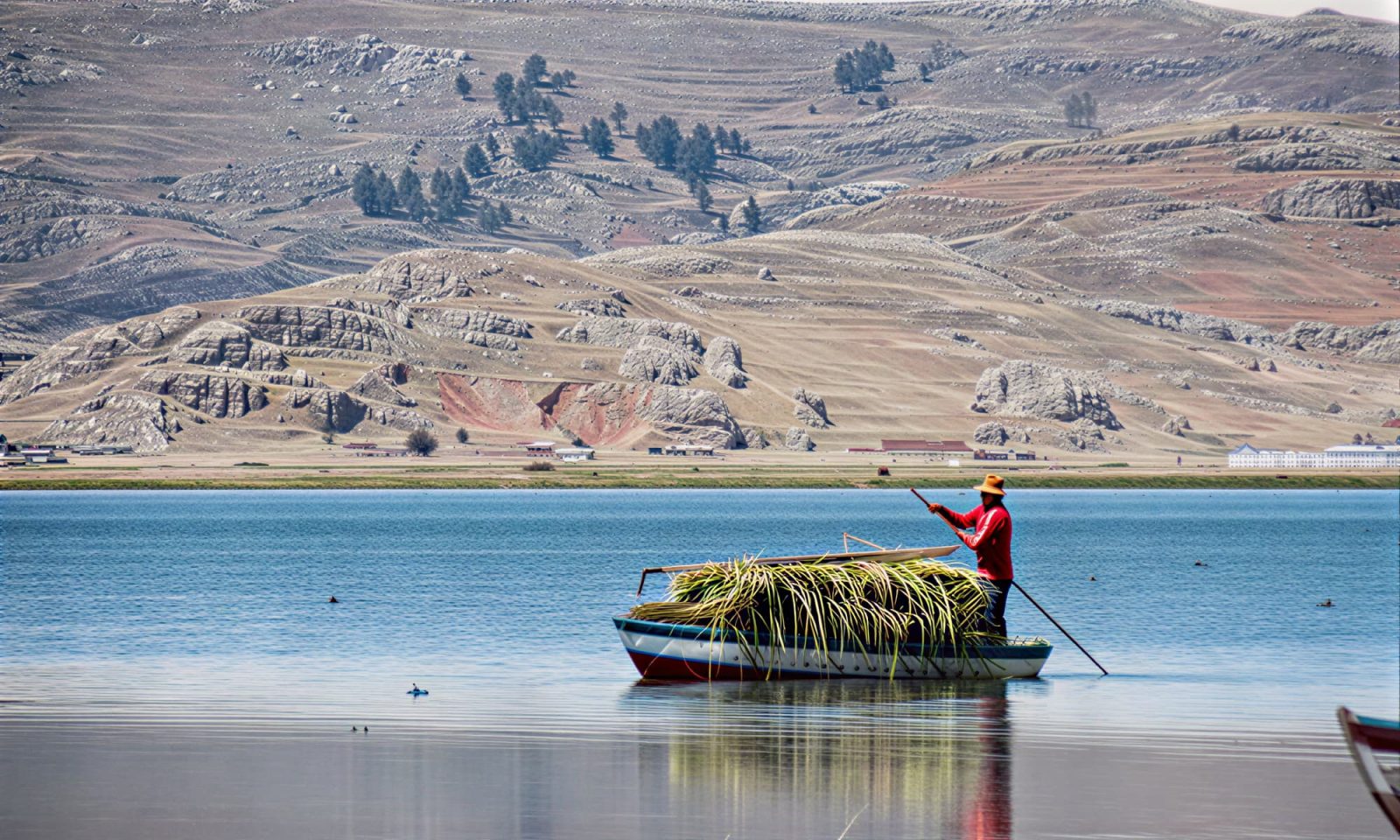 A Peruvian man uses a pole to push his boat through the water with mountains in the background