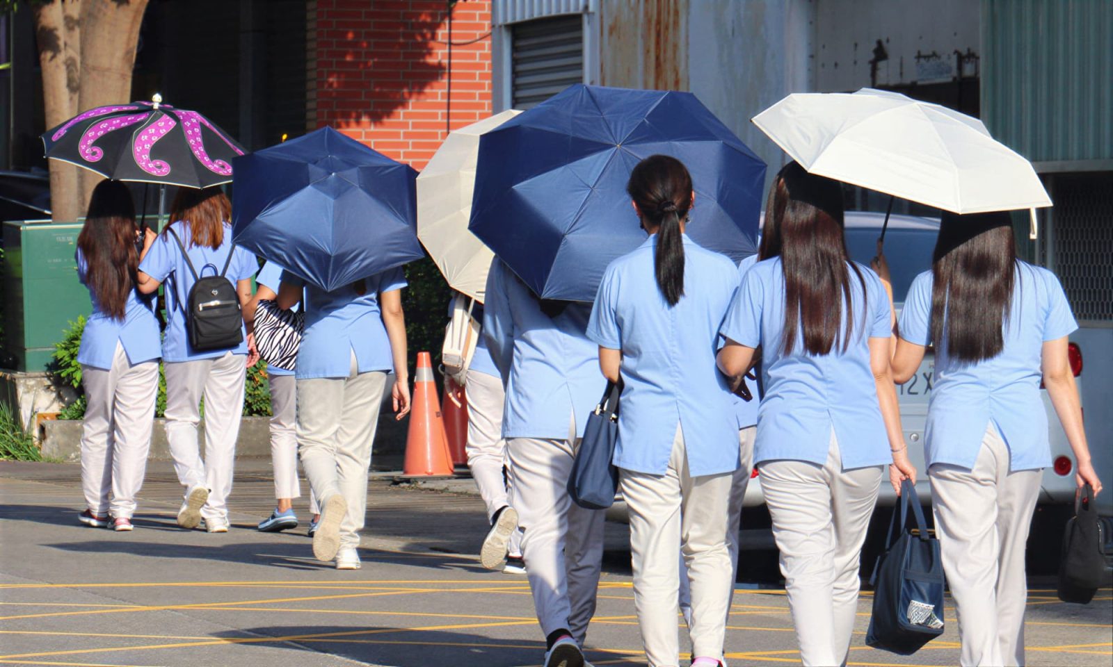 A group of women dressed in blue and white uniforms are walking with umbrellas towards buildings